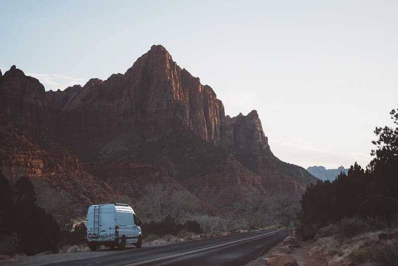 White sprinter van with a ladder and stickers parked in front of a scenic view in Zion National Park during sunset